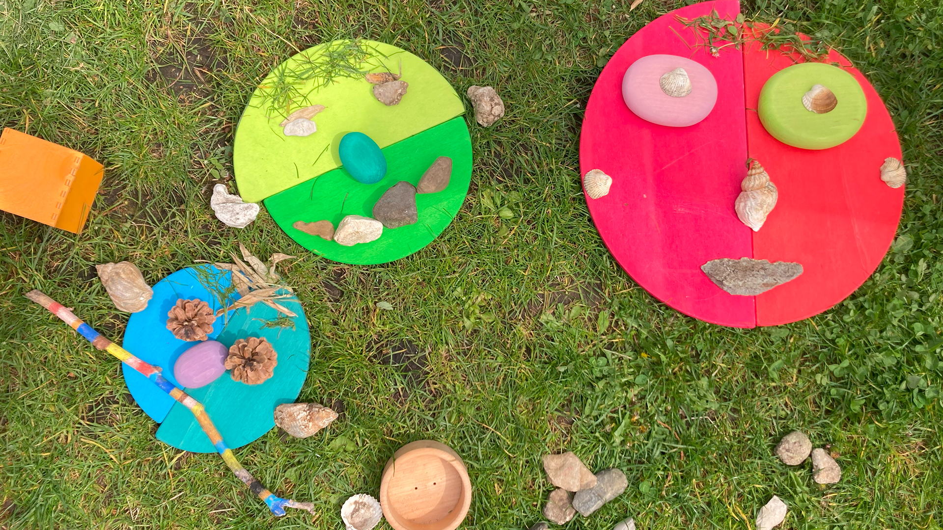 Bunte Gesichter aus Holzbausteinen und Naturmaterialien liegen im Gras.